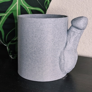 Könnte beinhalten: Ein grauer Keramik-Pflanztopf in Form einer Hand mit herausragendem Daumen. Der Topf steht auf einer schwarzen Oberfläche.