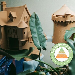 Puede incluir: Castillo y torre en miniatura impresos en 3D de color marrón claro. El castillo tiene múltiples ventanas, una chimenea y una pequeña torreta. La torre tiene un techo cónico y un cuerpo cilíndrico. El logotipo Happy Place 46 es visible.