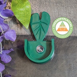 Könnte beinhalten: Ein grüner, 3D-gedruckter Handyständer in Form eines Blattes mit einem kleinen Loch in der Mitte. Der Ständer hat ein Logo an der Seite, auf dem "The Happy Place 46 3D Prints Factory" steht.
