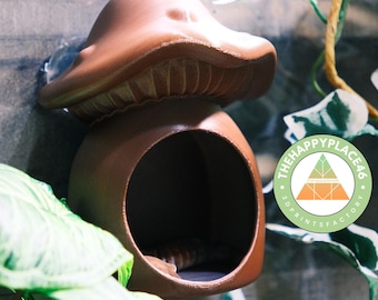 Può includere: Un nascondiglio in ceramica marrone a forma di fungo con un'apertura rotonda, adatto per piccoli rettili o anfibi. Il nascondiglio è montato su una parete e ha un adesivo con il testo "The Happy Place 46 3D Print Factory".