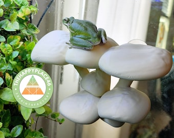 Peut inclure: Une grenouille verte arboricole perchée sur une plateforme en forme de champignon blanc. La plateforme fait partie d'une structure de champignons à plusieurs niveaux. L'arrière-plan présente une plante verte feuillue et un logo qui dit "The Happy Place 46 3D Print Factory".