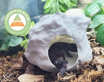 Puede incluir: Una escondite para reptiles impresa en 3D de color blanco con una superficie texturizada. Un pequeño gecko está dentro del escondite. El escondite está etiquetado como "The Happy Place 46 3D Prints Factory".