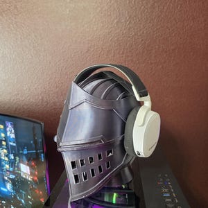 May include: A dark gray, 3D-printed knight helmet with a pointed visor and rectangular eye slits. The helmet is topped with a pair of white and black over-ear headphones featuring a padded headband and circular ear cups with visible branding.