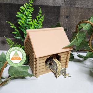 May include: A light brown log cabin-style reptile hide with a light pink roof. A crested gecko is partially visible inside the hide.