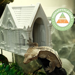 Puede incluir: Una iglesia en miniatura gris impresa en 3D con puertas y ventanas arqueadas. Un gecko crestado se posa en una rama negra frente a la iglesia. Los logotipos de The Happy Place 46 y 3D Prints Factory son visibles.