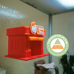Puede incluir: Modelo en miniatura rojo y naranja de un restaurante drive-thru. El modelo presenta un letrero que dice "Drive Thru" y un logotipo. Es un artículo impreso en 3D, posiblemente un juguete o decoración. El logotipo de The Happy Place 46 es visible.