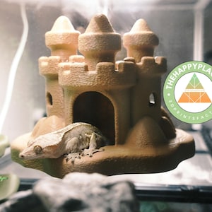Peut inclure: Un refuge pour reptile en forme de château de couleur beige avec trois tourelles et des portes arquées. Un gecko à crête se repose à l'intérieur du refuge. Le logo The Happy Place 46 est visible.