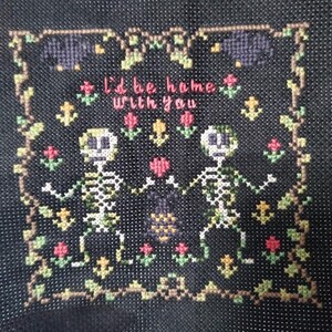 Op de afbeelding: Kruissteekontwerp op zwarte stof met twee skeletten die elkaars hand vasthouden, omringd door bloemen- en rankmotieven. De tekst "I'd be home with you" is in roze gestikt. Het ontwerp bevat rode, gele en groene accenten.