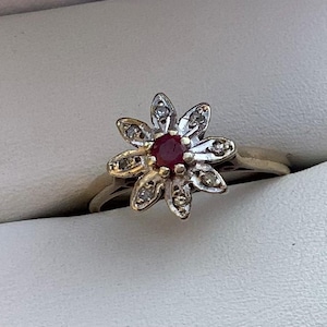Può includere: Anello d'oro con design a fiore, con una gemma rossa centrale e gemme chiare circostanti. Il design floreale dell'anello è posto su uno sfondo bianco, evidenziando i dettagli intricati.