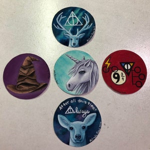 May include: Four round painted ceramic coasters with different Harry Potter themed designs. The first coaster features a white stag with antlers and a triangle with a circle inside. The second coaster features a brown wizard's hat on a purple background. The third coaster features a white unicorn with a blue horn on a blue and green background. The fourth coaster features a red background with a lightning bolt, a triangle with a circle inside, and the numbers 9 and 3/4.