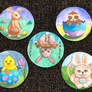 Puede incluir: Cinco adornos pintados al agua con temática de Pascua que representan un conejo sosteniendo una cesta de huevos, un perezoso en un huevo, un pollito en un huevo, un conejo sosteniendo un columpio y un gato con orejas de conejo sosteniendo un huevo.