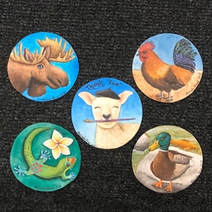 May include: Five round painted wooden ornaments with different animal designs. The ornaments feature a moose, a rooster, a lamb, a gecko, and a duck. Each ornament has the artist's name and the year painted on the bottom.
