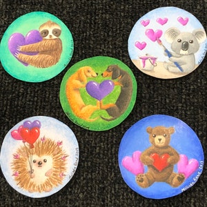 May include: Five round, colorful painted ornaments with various animal and heart designs. The ornaments feature a sloth holding a heart, a dachshund and a miniature dachshund holding a heart, a koala painting a heart, a hedgehog with balloons, and a bear holding a heart.