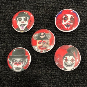 Puede incluir: Cinco botones redondos rojos y blancos con diferentes imágenes de payasos. Los botones tienen los nombres "Joker", "Cotton", "Teddy", "Karsten" y "Trick" escritos en ellos.