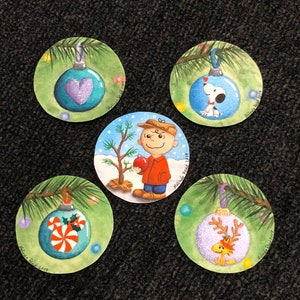 Könnte beinhalten: Fünf runde Ornamente mit bemalten Designs. Die Ornamente zeigen ein Herz, einen Hund, einen Weihnachtsbaum, einen Zuckerstock und ein Rentier. Jedes Ornament ist von einem grünen Tannenzweig umgeben und hat einen weißen Hintergrund.
