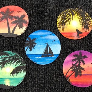Puede incluir: Cinco pinturas redondas de acuarela de puestas de sol tropicales con palmeras, un velero y una cola de ballena. Las pinturas están sobre una superficie gris oscura.