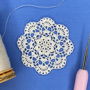 May include: A white, handmade crochet doily with a floral design, a spool of white cotton thread, and a pink-handled crochet hook, all on a blue fabric background. The thread spool reads "- 100% Cotton/Co".