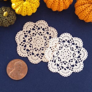May include: Two round, off-white crocheted doilies with intricate floral patterns and scalloped edges. A copper-colored United States penny is positioned next to the doilies to provide a clear sense of scale for the small, delicate textile items.