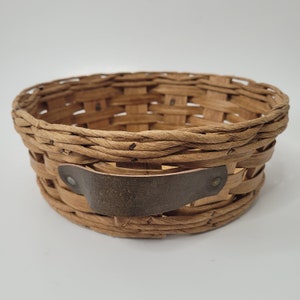 May include: A woven wicker basket with a brown leather handle. The basket is round and has a natural brown colour.