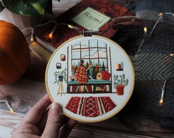 May include: Embroidered hoop art featuring a cozy scene of a person sitting by a window with a book, surrounded by plants and bookshelves. The scene is framed by a wooden hoop and includes a colorful rug.