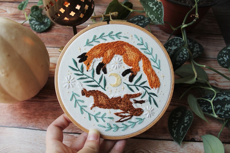 May include: Embroidered hoop art featuring a fox and a rabbit jumping in a whimsical forest scene with green leaves, white flowers, and a crescent moon.