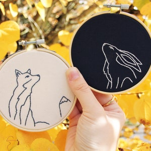 May include: Two embroidery hoops with black and white fabric. The hoop on the left has a white fabric with a black outline of a fox. The hoop on the right has a black fabric with a white outline of a rabbit.