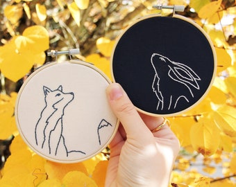 May include: Two embroidery hoops with black and white fabric. The hoop on the left has a white fabric with a black outline of a fox. The hoop on the right has a black fabric with a white outline of a rabbit.