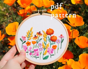 May include: A hand-stitched embroidery hoop featuring a colorful floral design. The embroidery includes various flowers in shades of pink, orange, yellow, and purple. The text "pdf pattern" is displayed above the embroidery. The background is filled with bright orange flowers.