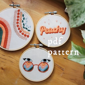 Puede incluir: Tres piezas de arte de bordado en aro con un arcoíris, un par de gafas y la palabra "Peachy" en una fuente retro. Los aros están sobre una superficie de madera.