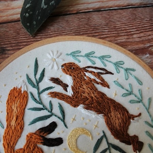 May include: Embroidered hoop art featuring a brown rabbit jumping up towards a white flower and a red fox with its tail curled up. The background is white with green leaves and a yellow crescent moon.