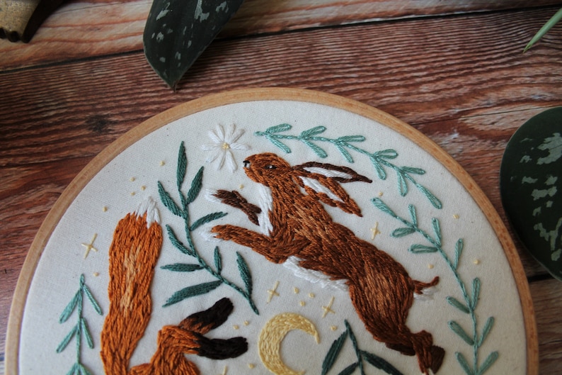 May include: Embroidered hoop art featuring a brown rabbit jumping up towards a white flower and a red fox with its tail curled up. The background is white with green leaves and a yellow crescent moon.