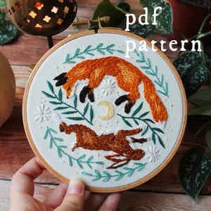 May include: Embroidered hoop art featuring a fox and a rabbit jumping in the air, surrounded by green leaves and white flowers. The hoop is made of wood and has a white fabric background. The text "pdf pattern" is written in black letters at the top of the image.