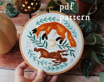 May include: Embroidered hoop art featuring a fox and a rabbit jumping in the air, surrounded by green leaves and white flowers. The hoop is made of wood and has a white fabric background. The text "pdf pattern" is written in black letters at the top of the image.