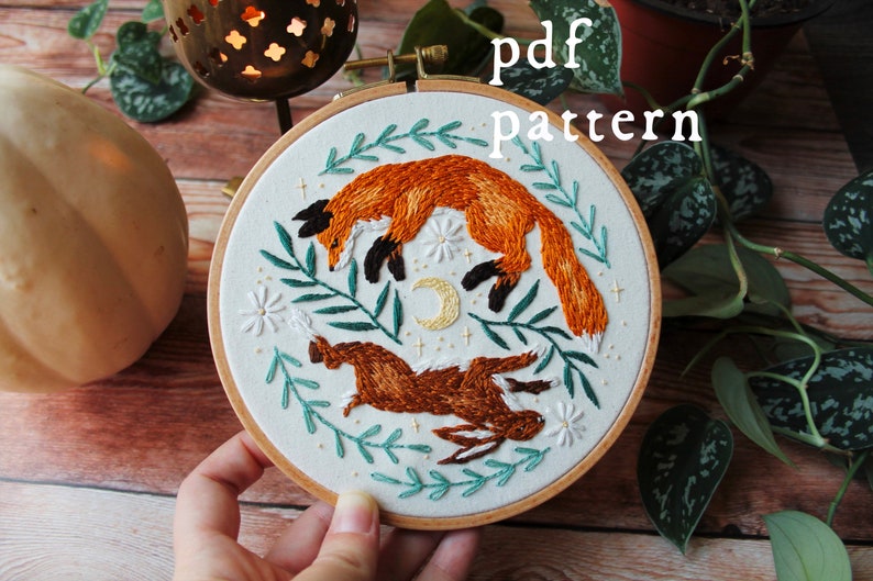 May include: Embroidered hoop art featuring a fox and a rabbit jumping in the air, surrounded by green leaves and white flowers. The hoop is made of wood and has a white fabric background. The text "pdf pattern" is written in black letters at the top of the image.