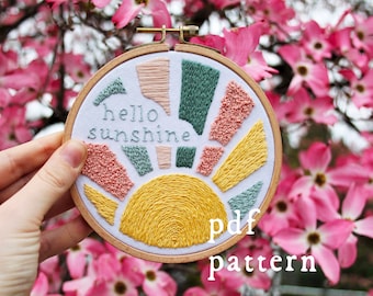 Könnte beinhalten: Stickrahmen-Kunst mit einer bunten Sonnen-Stickerei und dem Text "hello sunshine". Der Rahmen wird in einer Hand vor einem Hintergrund aus rosa Blüten gehalten. Der Text "pdf pattern" steht unter dem Rahmen.