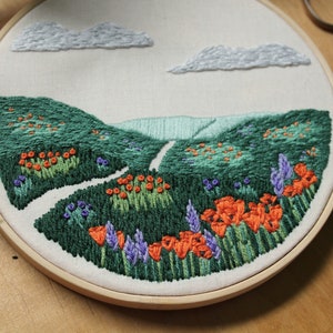 May include: Embroidered hoop art depicting a landscape with green hills, a winding path, and orange flowers. The background features a light blue sky with white clouds.