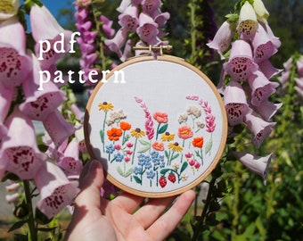 May include: A close-up of a hand holding a white embroidery hoop with a colourful floral design. The hoop is surrounded by pink foxglove flowers. The text "pdf pattern" is written above the hoop.