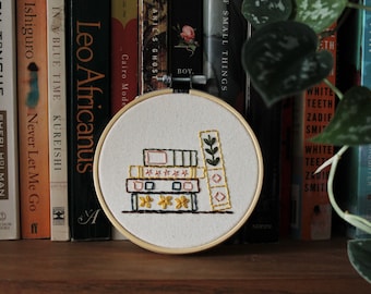 以下が含まれることがあります： 手縫いの刺繍枠のアップで、本と葉の植物が積み重ねられています。本は茶色、黄色、赤の色合いで刺繍されています。植物は緑色で刺繍されています。枠は木の棚にあり、後ろには本が並んでいます。