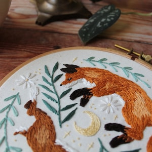 May include: Embroidered hoop art featuring two foxes, one brown and one orange, with green leaves and white flowers. The foxes are surrounded by a crescent moon and stars.