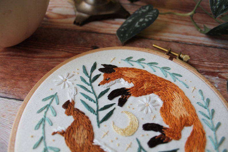 May include: Embroidered hoop art featuring two foxes, one brown and one orange, with green leaves and white flowers. The foxes are surrounded by a crescent moon and stars.