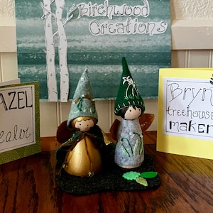May include: Two handmade fairy dolls with felt hats and wings, standing on a green felt base. A green card with the text "Hazel Healer" and a yellow card with the text "Bryn treehouse maker" are also visible. The background features a sign that says "Birchwood Creations".