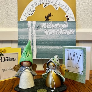 May include: Two handmade fairy dolls with felt hats and wings sit on a dark gray felt base. A sign reads "Welcome to the World of Birchwood Creations." Two cards read "Willow herbalist" and "Ivy Gatherer."