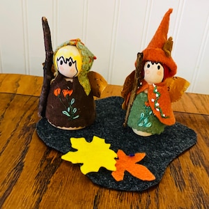 May include: Two handmade felt fairy dolls with wooden stick staffs. One doll wears a brown dress with orange flower embroidery and a green and yellow head covering. The other doll wears a green dress and orange cape with white dot accents and an orange hat. Felt leaves are on the base.