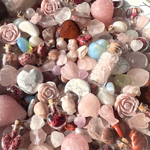 May include: A collection of rose quartz crystals, carved roses, hearts, and other shapes. Small glass bottles filled with rose petals and crystals are also present. The color palette is soft pinks, whites, and reds.