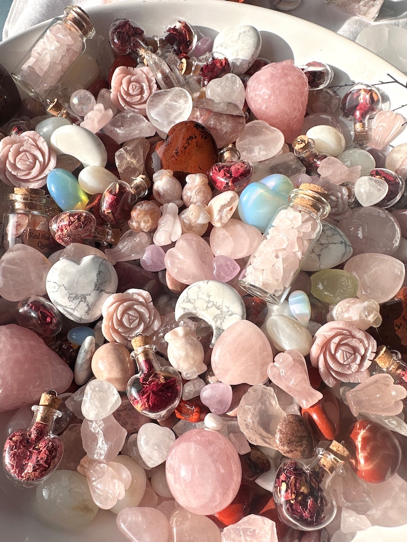 May include: A collection of rose quartz crystals, carved roses, hearts, and other shapes. Small glass bottles filled with rose petals and crystals are also present. The color palette is soft pinks, whites, and reds.