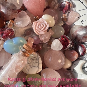 May include: A collection of various pink and clear crystals, including heart-shaped stones, spheres, and a carved rose. Small glass bottles with cork stoppers are also visible, some filled with dried rose petals. The text in the image reads "Roses & Strawberries are more costly, so they are only available as an add on item."