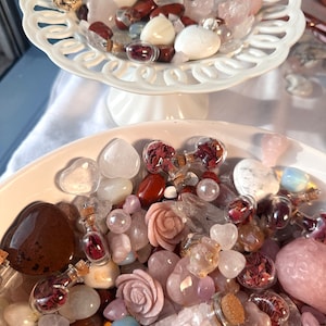 May include: A collection of various gemstones and crystals in shades of pink, white, and brown, displayed in white, decorative dishes. The assortment includes heart-shaped stones, carved roses, and small glass bottles filled with dried petals.