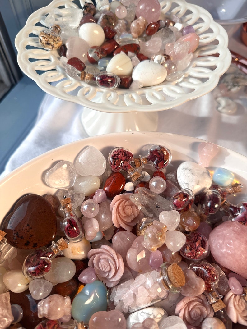 May include: A collection of various gemstones and crystals in shades of pink, white, and brown, displayed in white, decorative dishes. The assortment includes heart-shaped stones, carved roses, and small glass bottles filled with dried petals.