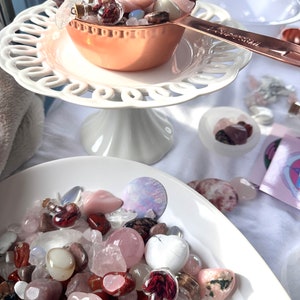 May include: A collection of crystals and gemstones in various shapes and colors, including rose quartz, displayed in a copper measuring cup and white bowls. A white, tiered cake stand holds the cup. Small glass vials with corks are also visible.