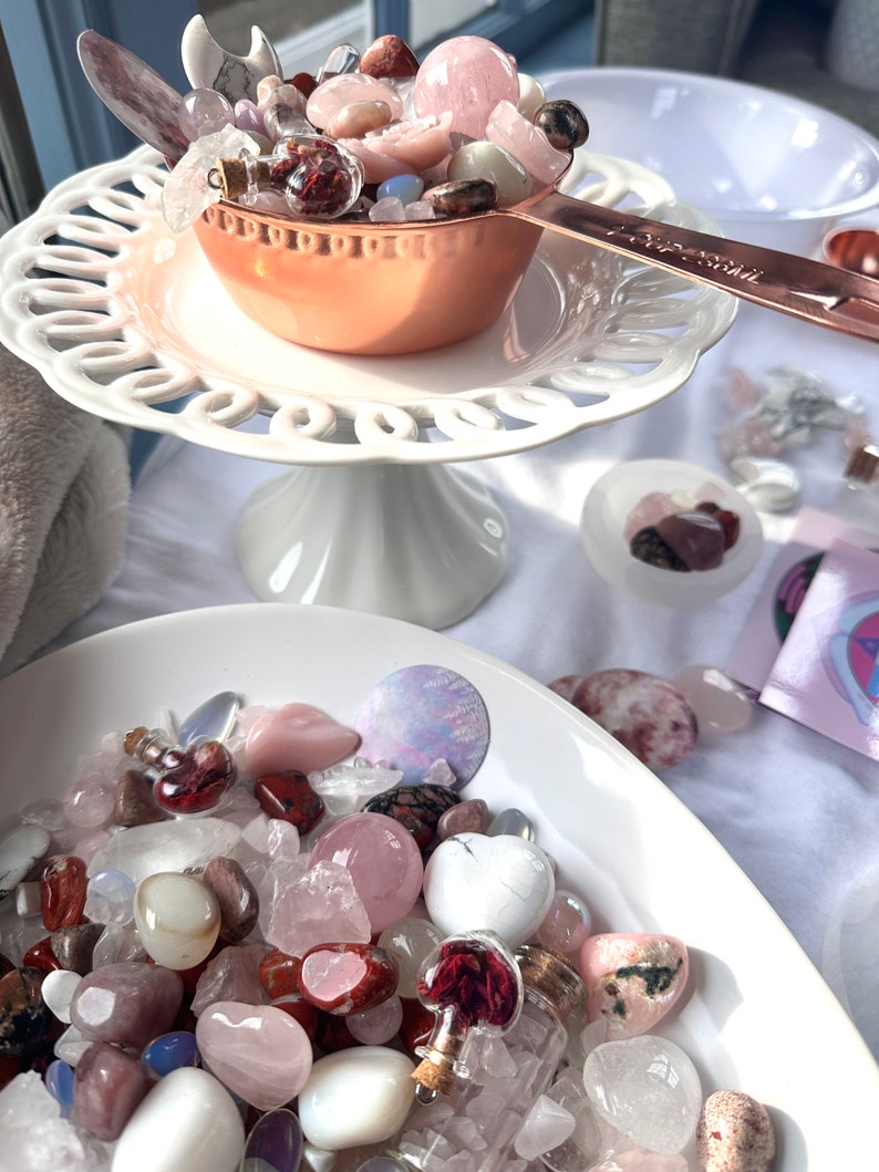 May include: A collection of crystals and gemstones in various shapes and colors, including rose quartz, displayed in a copper measuring cup and white bowls. A white, tiered cake stand holds the cup. Small glass vials with corks are also visible.